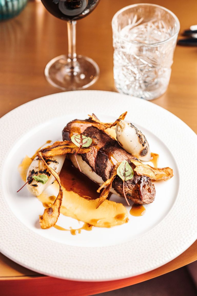 A dish displayed with four slices of tender beef, with charred shallots and garnish