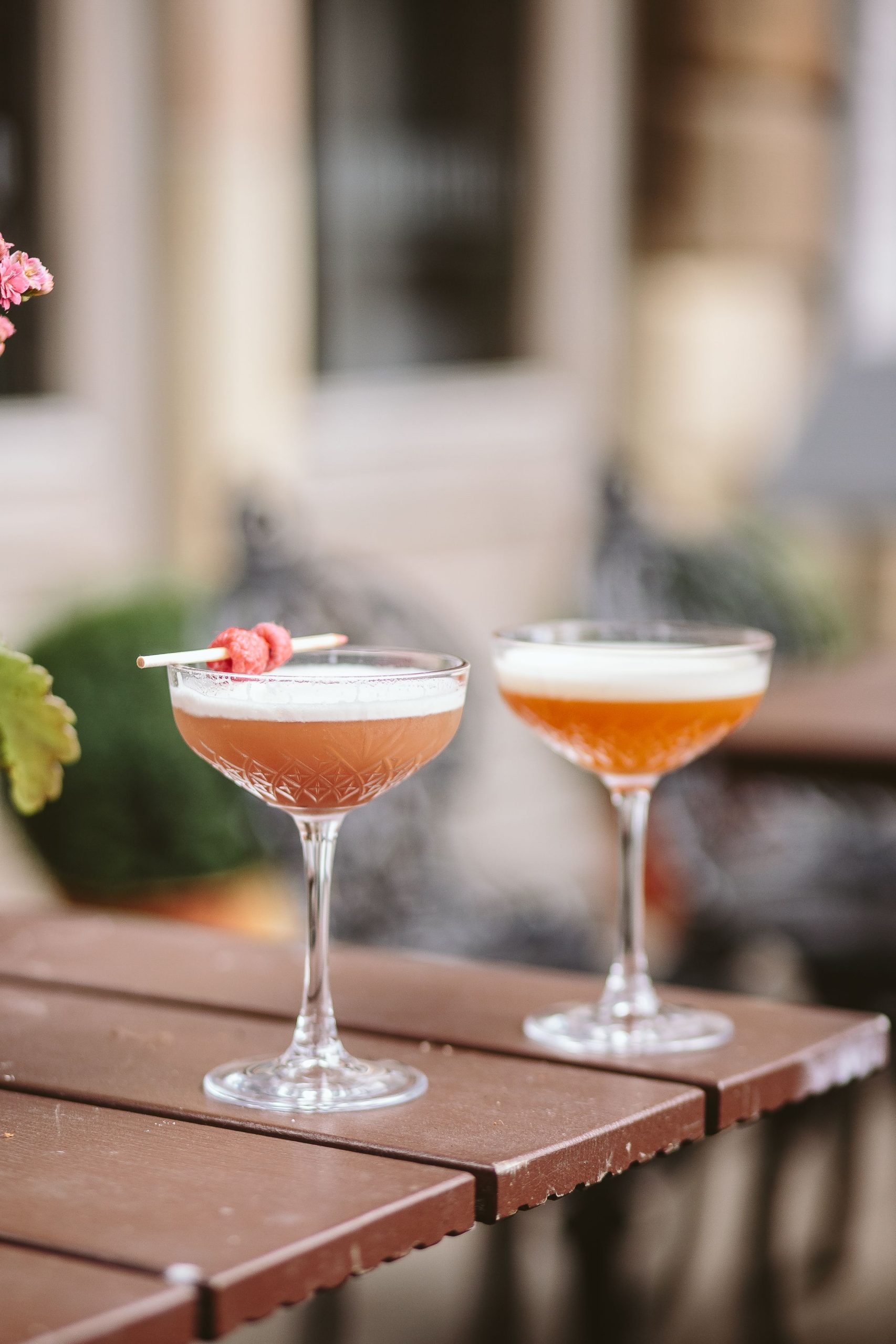Two cocktails, pink and orange in coupe glasses outside on a wooden table